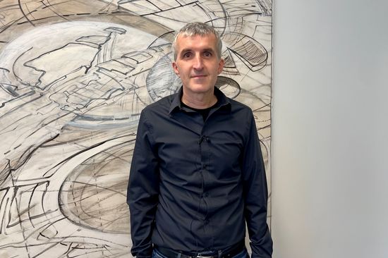 Halb-Porträt Johannes Böhm stehend vor einer Wand mit abstraktem Gemälde in braun-grün