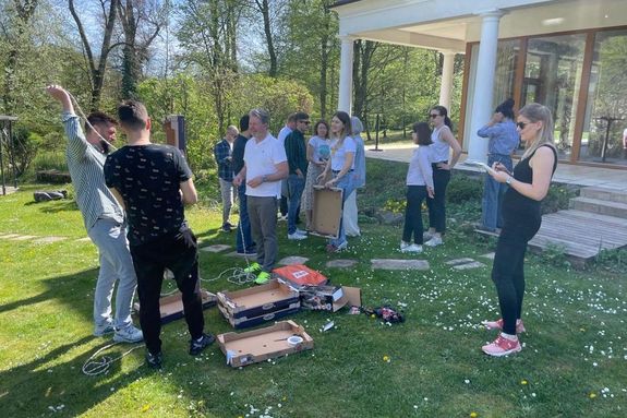 Außenaufnahme: Studierende stehend auf grüner Wiese, Schachteln am Boden