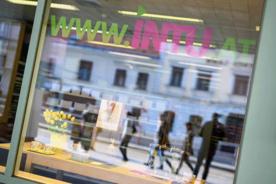 Außenfenster des INTU-Shops, in der sich der sich die Fassade des gegenüberliegenden Hauses und die Passant_innen spiegeln.