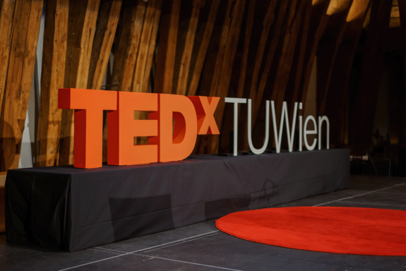 TEDx in orange TUWien in weißen Lettern auf einem schwarzen Sockel. Roter runder Teppich im Vordergrund. Im Hintergrund die Planken des Kuppelsaals.