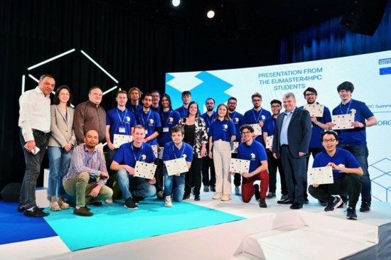 Gruppenbild von Studierenden in blauen T-Shirts, die Urkunden halten. Dunkler Hintergrund und großer Screen.
