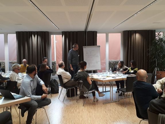 [Translate to English:] Group of people sitting in meeting room, one person at flipchart