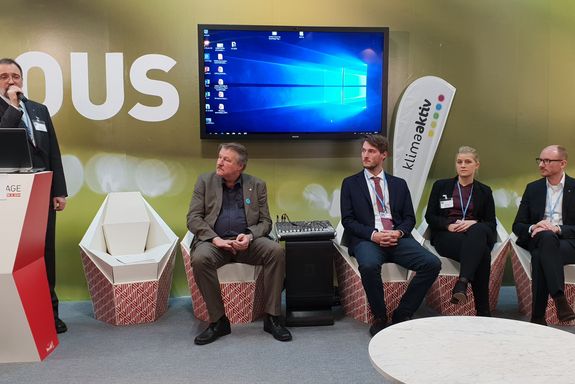 (from left to right) Dipl.-Ing. Peter Heimerl moderating, standing. four presenters sitting, watching him: Carl W. Seitz (Kohlbach Energy Systems GmbH), Moritz Bühner (Fritz Egger GmbH), Dr. Simone Spitzer (TU Wien), Prof. Dr. René Hofmann (TU Wien) - all in front of a big flat screen