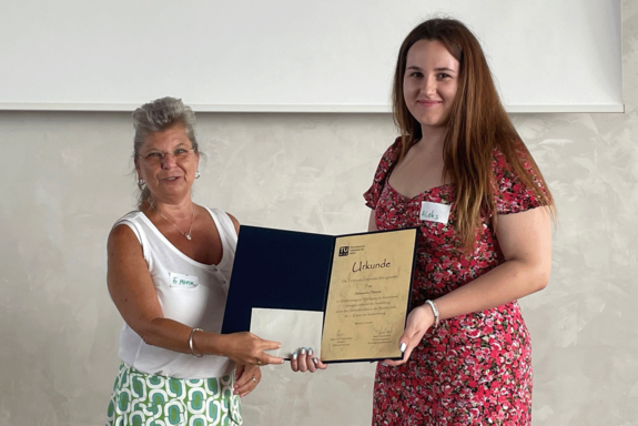 Manuela Marik in weißem, ärmellosen T-Shirt und grün-weißem Rock präsentiert gemeinsam mit Aleksandra Hosovic die Urkunde. Hosovic trägt ein rotes, gemustertes Sommerkleid.
