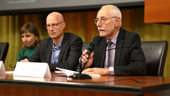 Three speakers, one female, two male, sitting at the front desk