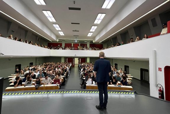 Blick von Bühne mit Moderator mit Rücken zu Kamera in Hörsaal mit sitzend Studierenden
