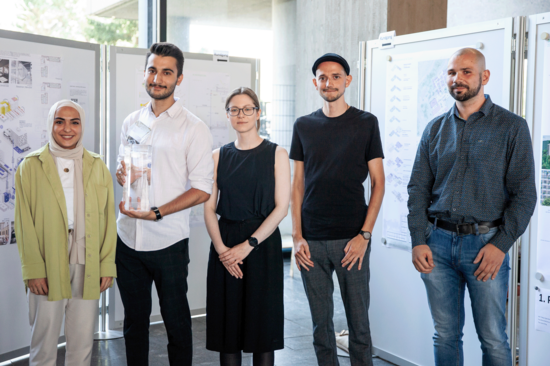 Gruppenfoto mit fünf Teilnehmer_innen der Siegerteams. Im Hintergrund die Präsentation der Entwürfe.