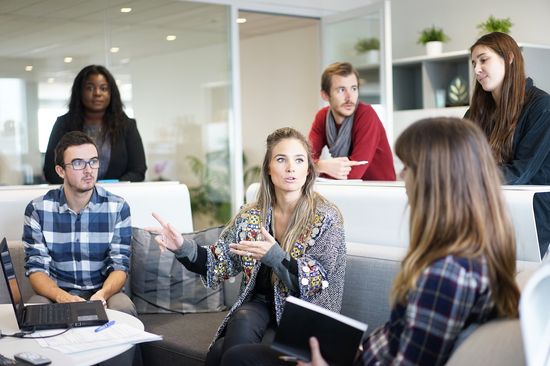 A group of six people of different genders are sitting and standing in a modern office. They are engaged in lively discussion.