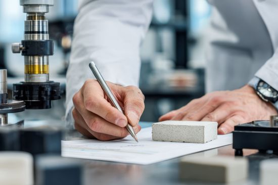 Die Hände eines Werkstoffingenieurs, der Daten erfasst, während er ein zerbrochenes Keramikstück untersucht, umgeben von Laborgeräten zur Festigkeitsanalyse und Validierung