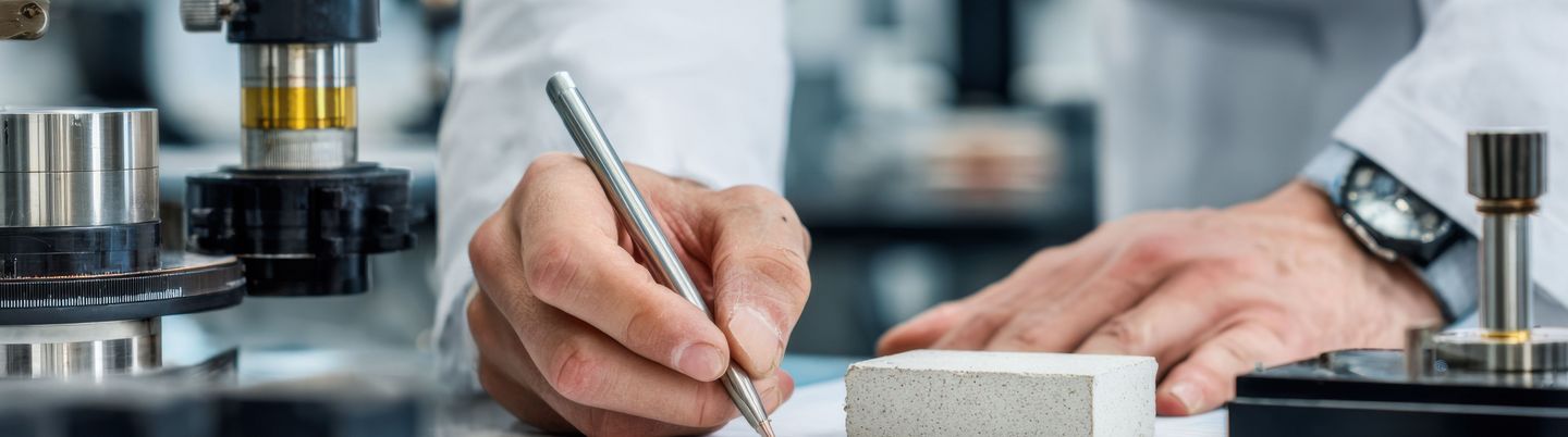 Die Hände eines Werkstoffingenieurs, der Daten erfasst, während er ein zerbrochenes Keramikstück untersucht, umgeben von Laborgeräten zur Festigkeitsanalyse und Validierung