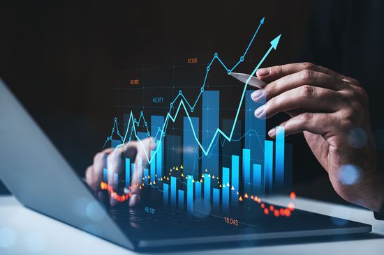 Geschäftsmann analysiert Finanzdaten-Diagramme auf einem Laptop mit einem Stylus in einem futuristischen Interface-Konzept auf dunklem Hintergrund mit leuchtenden Grafiken und Balken.