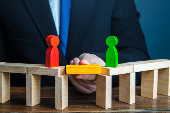 Two bridges made of wooden blocks, each with a wooden figure standing on it. The two bridges are connected by a wooden block of a different colour.