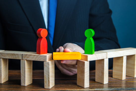 Zwei Brücken aus Holzklötzen, auf denen jeweils eine Holzfigur steht. Die beiden Brücken werden von einem andersfarbigen Holzblock verbunden.