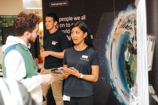 Ein Mann und eine Frau unterhalten sich auf einer Messe. Im Hintergrund sieht man einen weiteren Mann im Gespräch sowie ein großes Rollup einer Firma.