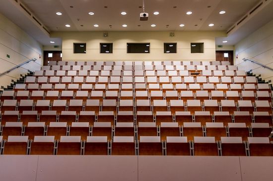 An empty lecture hall with tiered seating. A projector hangs from the ceiling. Shot from the perspective of the lecturer.