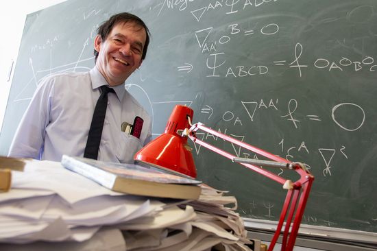 Mann hinter auf Schreibtisch aufgestapeltem Papier, vor beschriebener Tafel