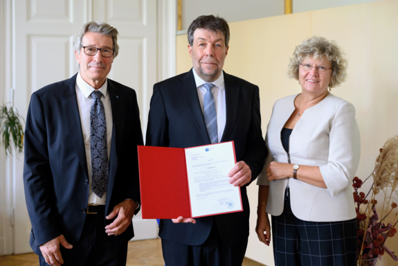 Honorarprofessor Peter Schöggl präsentiert seine Urkunde, links von ihm Vizerektor Matyas, rechts von ihm Rektorin Seidler. Alle sind festlich gekleidet.