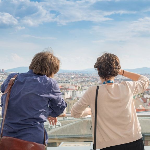 Zwei Menschen sind von hinten zu sehen. Sie stehen auf dem Dach eines Gebäudes und blicken auf Wien.