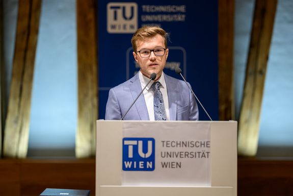 junger mann mit Brille und hellblauem Anzug hinter Pult mit Mikro