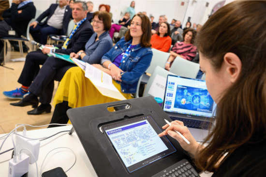 Im Vordergrund des Bildes eine Präsentation auf einem Tablet, rechts dahinter ein Laptop und schräg verlaufend dahinter Pulbikumsreihen. In der ersten Reihe Mtglieder des Rektorats der TU Wien.