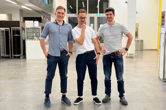 Drei junge männliche Personen stehen gemeinsam in einem großen Raum, sie lächeln in die Kamera.