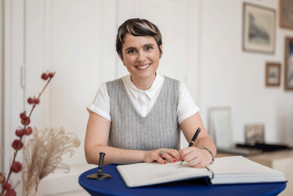 Teresa Liberto beim Unterschreiben der Urkunde. Lächelt in die Kamera, trägt einen grau-beigen Pollunder über weißem Shirt.