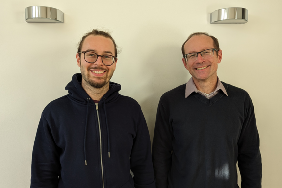 Das Foto zeigt Valentin und Derek, die nebeneinander vor einer weißen Wand mit zwei Wandleuchten stehen.