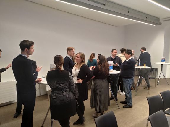 Alumni, Studierende und Vortragende beim persönlichen Austausch bei Snacks und Getränken (an Stehtischen) im Anschluss an den Vortrag