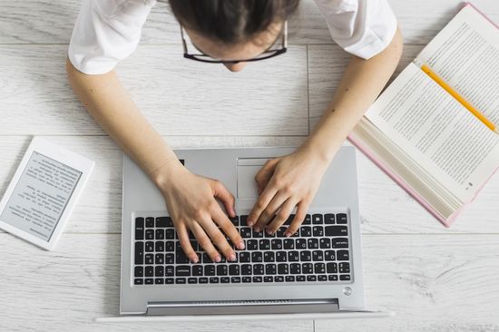 Aufsicht: Frau liegend, tippt auf Laptoptastatur, neben ihr ein aufgeschlagenes Buch