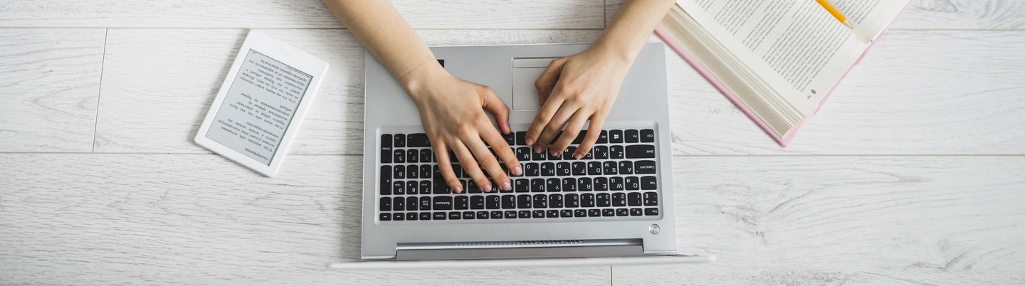 Aufsicht: Frau liegend, tippt auf Laptoptastatur, neben ihr ein aufgeschlagenes Buch