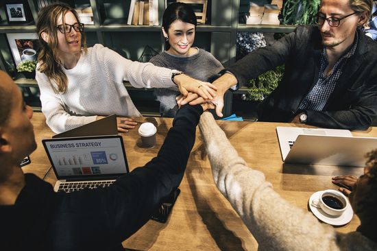 Junge bzw Mensch mittleren Alters mit ihren Laptops um einen Holztisch. Sie legen die Hände in der Mitte übereinander als ob sie diese gleich hochwerfen werden.