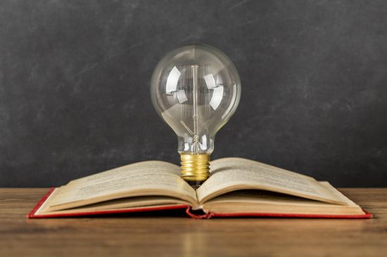 A large light bulb stands between the pages of an open book. The book lies on a wooden surface and has a red cover.