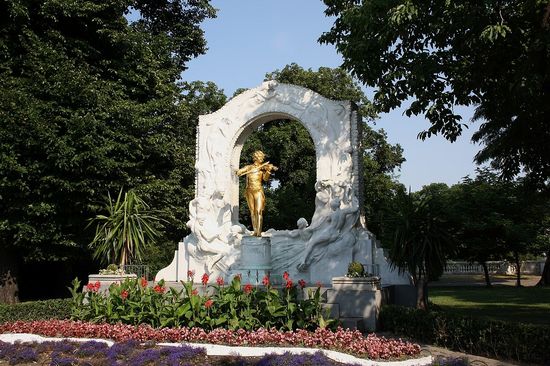 Johann-Strauß-Denkmal im Wiener Stadtpark: Das vergoldete Bronzestandbild auf einem Marmorsockel zeigt den Wiener Walzerkönig stehend – ohne den dichten Backenbart – mit einer Geige in der Hand vor einem mit Blättern und schwebenden Paaren reliefierten Laaser Marmorbogen.
