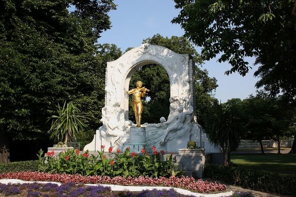 Johann-Strauß-Denkmal im Wiener Stadtpark: Das vergoldete Bronzestandbild auf einem Marmorsockel zeigt den Wiener Walzerkönig stehend – ohne den dichten Backenbart – mit einer Geige in der Hand vor einem mit Blättern und schwebenden Paaren reliefierten Laaser Marmorbogen.