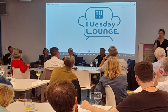 Ein Foto, auf dem man im Vordergrund das Publikum sieht und im Hintergrund eine Rednerin, eine Präsentation mit dem TUesday Lounge-Logo und Diskussionsteilnehmer_innen.