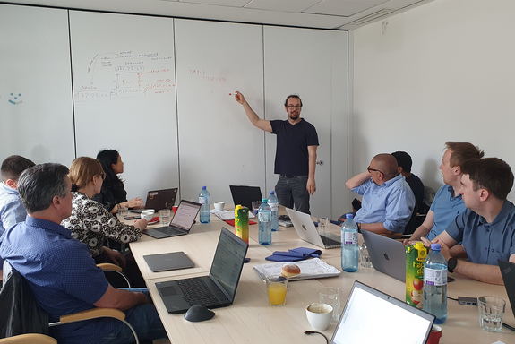 Das Foto zeigt mehrere Personen, die mit ihren Laptops an einem großen Tisch sitzen und zu einer Person schauen, die vor dem Tisch steht und an einer Wand Notizen macht.