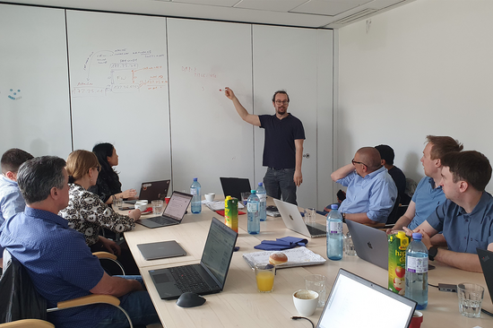 Das Foto zeigt mehrere Personen, die mit ihren Laptops an einem großen Tisch sitzen und zu einer Person schauen, die vor dem Tisch steht und an einer Wand Notizen macht.