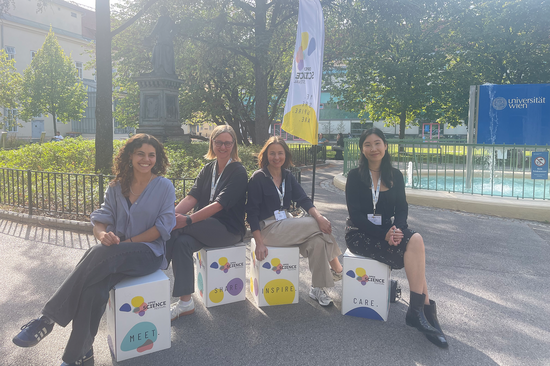 Four women sit in front of a green area and a fountain on cardboard boxes in the Open Science Festival design.