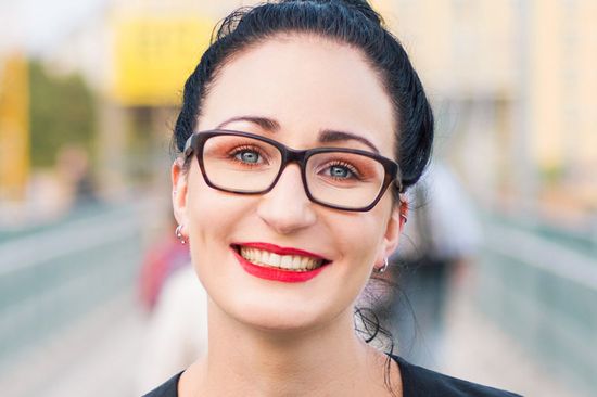 Portrait der lächelnden Miriam Unterlass: dunkle nach hinten gebundene glatte Haare, große schwarzumrandete Brille, kräftig rot geschminkte Lippen, das Augenmakeup ebenso im rötlichen Ton.