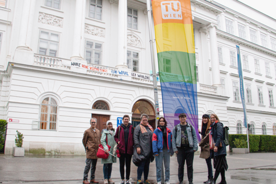 Gruppe von Personen vor dem TUW-Hauptgebäude bei der gehissten Regenbogenflagge.