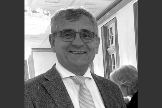 Schwarz-weiß-Porträt von Prof. Bucher mit dunklem Sakko, weißem Hemd und heller Krawatte in festlichem Saal, Kronleuchter links im Hintergrund.