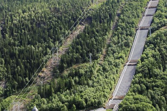 Ein Blick auf eine große Rohrleitung, die sich über eine bewaldete, hügelige Landschaft erstreckt. Die Rohre verlaufen parallel zueinander und sind in die grüne Umgebung eingebettet, während im Hintergrund Stromleitungen durch den Wald führen. Die Szene vermittelt eine Verbindung zwischen Industrie und Natur.
