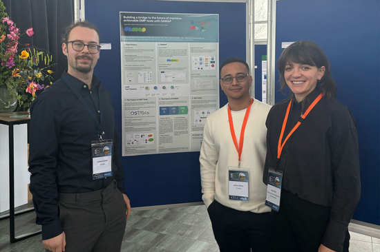 Zwei Männer und eine Frau stehen vor einer Posterwand mit einem Poster.