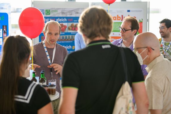 Wissenschaftler steht vor Posterwand und erklärt mehreren Besucher_innen seine Forschung.