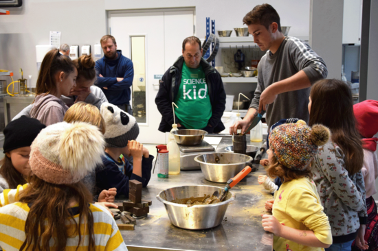 Kinder bei ihrem Workshop im Labor an einem Arbeitstisch mit Metallbehältern. Die Kinder im Vordergrund tragen gelb-weiß gestreifte Pullover, eines eine Kappe, eines eine Haube.