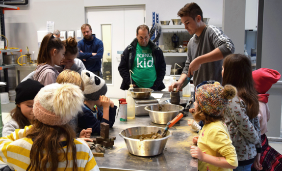 Kinder bei ihrem Workshop im Labor an einem Arbeitstisch mit Metallbehältern. Die Kinder im Vordergrund tragen gelb-weiß gestreifte Pullover, eines eine Kappe, eines eine Haube.