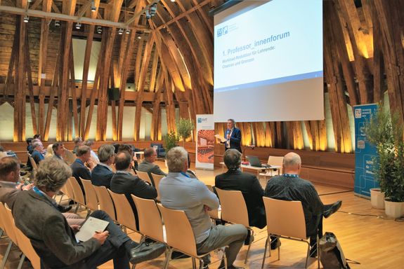 Kurt Matyas, Vizerektor Studium und Lehre, eröffnet das erste Professor_innenforum.  Publikum sitzen, Kurt Matyas stehend mit Mikrofon, über ihm eine Leinwand