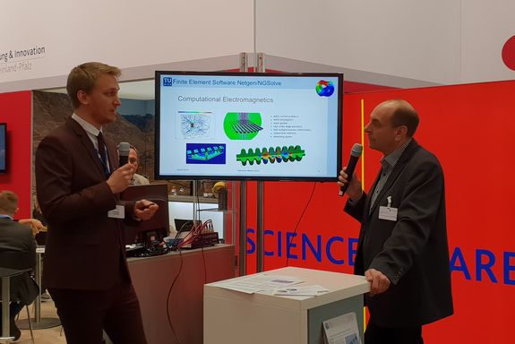 Christoph Lackner (Vienna University of Technology) in dialogue with the moderator of "Science Square" with microphones and a flatscreen in the back showing details on Netgen/NGSolve software.