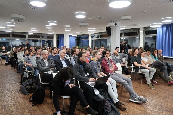 Blick auf das Publikum: zehn Reihen zu je zehn Sitzplätzen. Freie Plätze sind keine zu sehen. Im Hintergrund steht ein Kameramann und filmt.