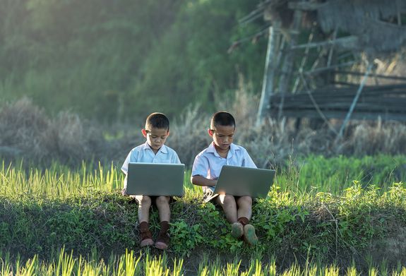 Zwei Buben sitzen konzentriert mit Laptops in einer grünen Landschaft, im Hintergrund Wald und die Reste eines Holzhauses mit Palmendach.
